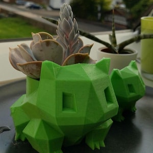 May include: Two bright green, low-poly, geometric planters shaped like a creature with pointed ears and square eye sockets. One planter holds a succulent plant with thick, pale purple-tinted leaves and a central flower spike growing upward.