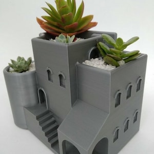Pode incluir: Um vaso cinzento impresso em 3D com a forma de um edifício de vários níveis com janelas em arco, uma escadaria e uma torre lateral. A estrutura apresenta três secções de plantação distintas preenchidas com gravilha branca e várias plantas suculentas verdes.
