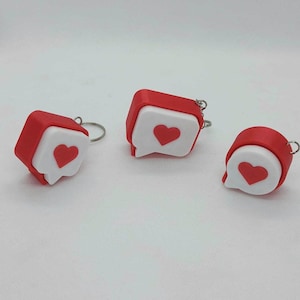 Peut inclure: Trois porte-clés en forme de bulles de dialogue avec une bordure rouge et un centre blanc arborant une icône de cœur rouge. Chaque porte-clés comprend un anneau en métal argenté fixé sur le dessus, présentant un design uniforme.