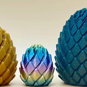 May include: Three egg-shaped objects with a textured, scale-like surface. The left egg is gold, the middle egg features a gradient of blue, purple, and yellow, and the right egg is a deep teal. Each object has a repeating diamond pattern.