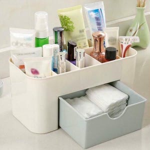 May include: A white plastic cosmetic organizer with multiple compartments and a pull-out drawer. The organizer holds various beauty products, including bottles, tubes, and small containers. The drawer contains a stack of white cotton pads.