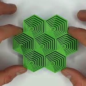 May include: A bright green, geometric puzzle composed of seven hexagonal shapes. Each hexagon features a nested spiral design, creating a 3D effect. The puzzle is held by hands, showcasing its size and form.