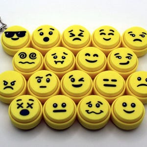 Könnte beinhalten: Eine Ansammlung von siebzehn runden gelben Knöpfen, von denen jeder ein anderes schwarzes Emoji-Gesicht mit verschiedenen Emotionen zeigt, angeordnet in einer Gruppe mit einem silbernen sternförmigen Metallschlüsselanhänger in der oberen linken Ecke.