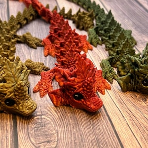 May include: A collection of articulated dragon figurines in various colors: olive green, red, and gold. The dragons have detailed scales, claws, and expressive faces with black eyes. They are arranged on a wooden surface.