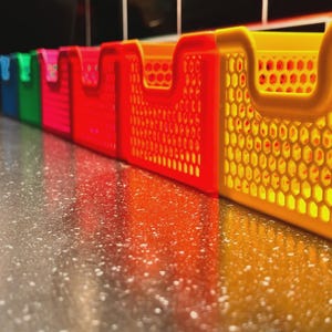 Könnte beinhalten: Eine Reihe bunter Kunststoff-Aufbewahrungsboxen in Blau, Grün, Pink, Rot und Gelb. Jede Box hat einen Griff und ein Wabenmuster an den Seiten. Die Boxen sind auf einer gesprenkelten Oberfläche angeordnet.