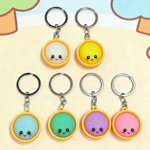 May include: Six round keychains featuring small, smiling dumpling characters in a yellow circular base. The dumplings come in white, yellow, light blue, green, purple, and pink, each with black eyes, a small mouth, and pink blush marks.