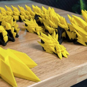 May include: A yellow and black articulated dragon toy, with a spiky, geometric design. The dragon is posed on a wooden surface, showcasing its segmented body and head. The toy is a vibrant yellow with black accents.