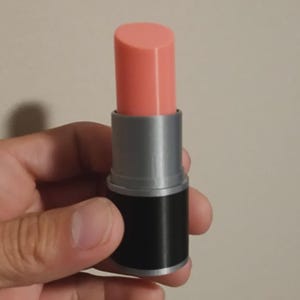 Könnte beinhalten: Ein zylindrischer Lippenstiftbehälter mit einem glatten, hellpfirsichfarbenen Kosmetikstift, der aus einem silberfarbenen Gehäuse herausragt und auf einem soliden schwarzen Sockel steht. Das Design ist schlicht und funktional und zeigt einen deutlichen Kontrast zwischen der weichen pfirsichfarbenen Spitze, dem metallisch silbernen Mittelteil und dem undurchsichtigen schwarzen Unterteil.