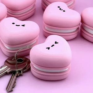 Peut inclure: Une collection de porte-clés macarons en forme de cœur rose avec des centres blancs et des expressions faciales noires. Chaque porte-clés présente une surface lisse et arrondie avec un anneau en métal attaché à l'une des pièces.