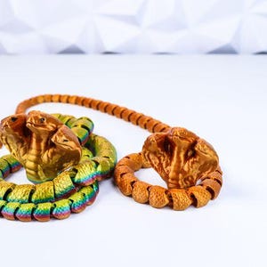 May include: Two decorative cobra sculptures. One has three heads and a rainbow-coloured body, the other has a single head and an orange body. Both have a textured, segmented design and are on a white surface.