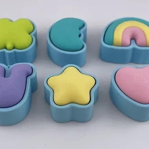 Op de afbeelding: Een set van zes lichtblauwe siliconen mallen, elk met een eigen vorm: een limoengroen klavertje, een groenblauwe halve maan, een regenboog, een paars hoefijzer, een gele ster en een roze hart, allemaal gerangschikt op een effen ondergrond.