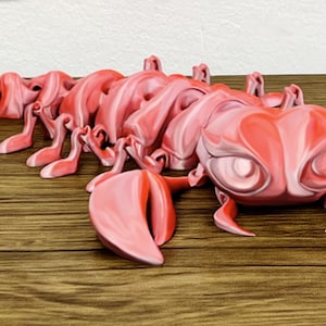May include: A decorative lobster figurine in shades of pink and red, with a cartoon-like aesthetic. The lobster is positioned on a wooden surface, highlighting its segmented body, claws, and detailed eyes. The figurine is a unique decorative item.