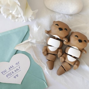 May include: Two brown otter figurines with white accents, arranged on white tissue paper. The otters are smiling and appear to be holding hands. A heart-shaped card with the text "YOU ARE MY OTTER HALF" is visible.