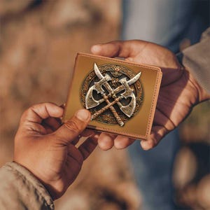 May include: A brown leather wallet featuring a detailed graphic of two crossed axes within a circular emblem. The axes have ornate silver blades and handles, set against a tan background. The wallet is held in two hands.