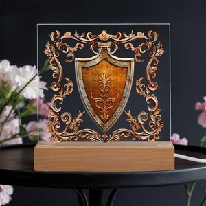 May include: A decorative lamp featuring a shield design. The shield is gold and silver, framed by ornate gold filigree on a clear acrylic panel. The lamp sits on a rectangular wooden base, with a power cord extending from the back. The background is dark.