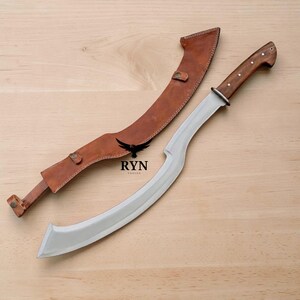 May include: A curved steel blade with a wooden handle featuring three metal rivets, accompanied by a matching brown leather sheath. The blade is marked with the text RYN FORGED and a black bird logo positioned between the sheath and the blade.