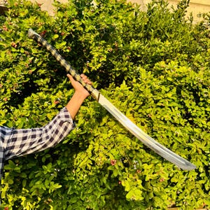 Pu&ograve; includere: Una lunga spada d'argento con un'impugnatura oro e nera &egrave; tenuta contro uno sfondo di fogliame verde. La lama &egrave; dritta e affilata, con un'elsa dorata e un'impugnatura nera. La spada &egrave; tenuta da una persona con una camicia a quadri.