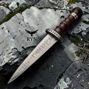 Könnte beinhalten: Ein handgefertigter Dolch mit einer gemusterten Stahlklinge und einem dunklen Holzgriff. Der Griff ist mit goldfarbenen Bändern verziert. Der Dolch liegt auf einer grauen Steinoberfläche. Der Text "RYN FORGED" und ein Vogellogo sind sichtbar.