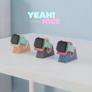Könnte beinhalten: Drei farbige Uhrenständer mit Smartwatches. Jeder Ständer hat eine andere Farbe: Pfirsich, Grau und Blau. Die Armbänder sind mintgrün. Im Hintergrund steht der Text "YEAH! NICE home decor".