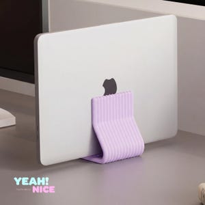 Könnte beinhalten: Ein silberner Laptop wird von einem hellvioletten Ständer auf einer grauen Oberfläche gehalten. Der Ständer hat ein gebogenes Design mit vertikalen Rippen. Der Text "YEAH! home decor NICE" befindet sich unten links.