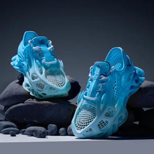 Può includere: Due paia di scarpe da ginnastica blu stampate in 3D con un design unico e intricato. Le scarpe sono appoggiate su rocce grigie.