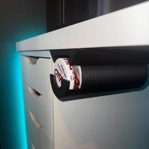 May include: A black 3D printed holder attached to the underside of a white desk, holding a can of Copenhagen Wintergreen Long Cut chewing tobacco.