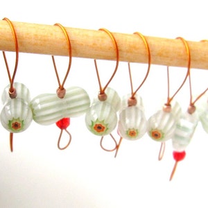 May include: Set of eight stitch markers with white and green striped beads and orange flower accents. The markers are hanging on a wooden dowel.