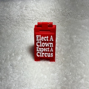 Può includere: Un cartello di plastica rosso con il testo "Elect A Clown Expect A Circus" in lettere bianche.