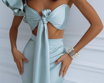 May include: A light blue satin two-piece set. The top is a knotted bandeau with a single ruffled sleeve. The matching skirt is a high-waisted, floor-length design. A pearl necklace and bracelet complete the look.