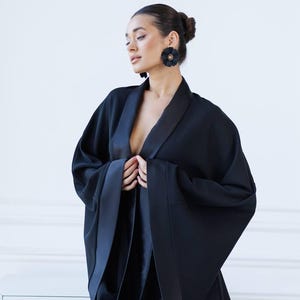 May include: A woman wearing a black satin kimono with a deep V-neckline and long sleeves. The kimono has a satin trim and is cinched at the waist. The woman is wearing black flower-shaped earrings.
