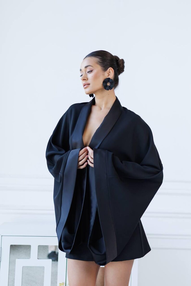 May include: A woman wearing a black satin kimono with a deep V-neckline and long sleeves. The kimono has a satin trim and is cinched at the waist. The woman is wearing black flower-shaped earrings.