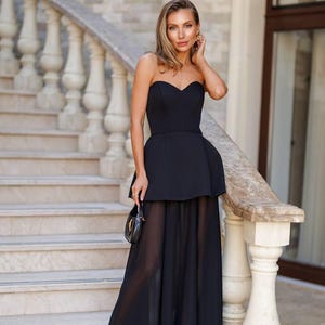 Puede incluir: Un vestido de noche negro sin tirantes con escote corazón y falda fluida y transparente. El vestido presenta un corpiño ajustado y un detalle de peplum en la cintura. La mujer lleva un pequeño bolso negro y sandalias negras. El fondo incluye una escalera de piedra y una barandilla de mármol.