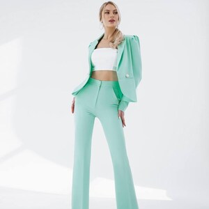 Puede incluir: Una mujer lleva un traje de pantal&oacute;n verde menta con un top corto blanco. El traje tiene una chaqueta con mangas abullonadas y cierre de bot&oacute;n. Los pantalones son de pierna ancha y tienen cintura alta.