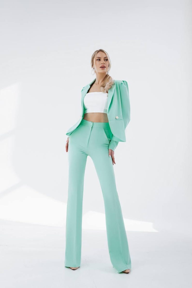 Puede incluir: Una mujer lleva un traje de pantal&oacute;n verde menta con un top corto blanco. El traje tiene una chaqueta con mangas abullonadas y cierre de bot&oacute;n. Los pantalones son de pierna ancha y tienen cintura alta.