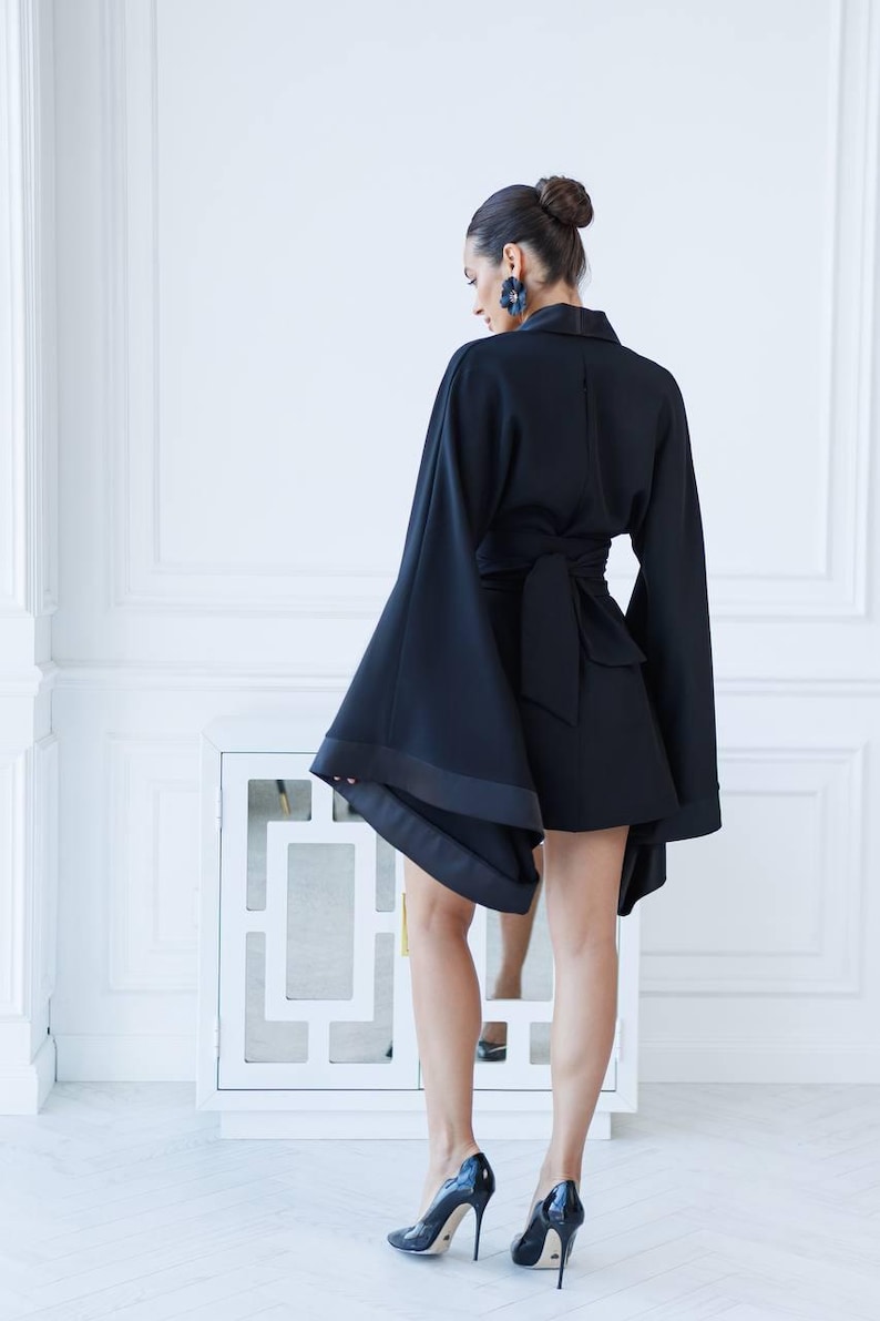 May include: A black short-sleeved dress with a wrap-style waist and wide, flowing sleeves. The dress is worn by a person with their back to the camera. The person is wearing black high heels.