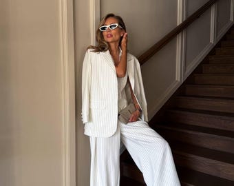 May include: A woman wearing a white pinstripe suit with a white blazer and wide-leg pants. She is standing on a staircase and wearing white sunglasses.