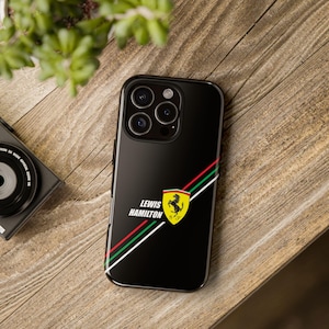 Może przedstawiać: Czarne etui na telefon z napisem "LEWIS HAMILTON" i żółtym logo Ferrari. Etui ma ukośny wzór w paski w kolorze czerwonym, zielonym i białym. Etui na telefon leży na drewnianej powierzchni.