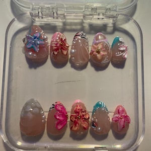 May include: A set of ten artificial nails in a clear plastic case. Each nail is a nude color with various floral designs in pink, blue, and white, accented with pearls and gold beads. The nails are oval-shaped.
