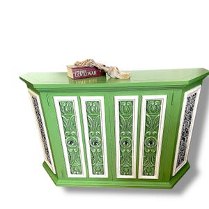 May include: A green cabinet with white trim and ornate carved details. The cabinet has four doors with decorative hardware and two side panels with a black and white pattern. A book titled "LOVE WAR" and a tassel are on top.