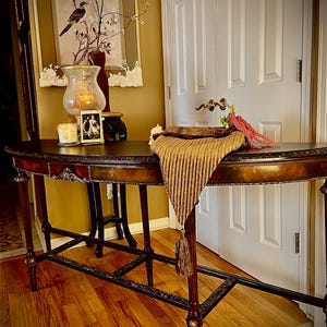 May include: A dark brown wooden console table with a curved top and intricate detailing. The table has two shelves and is decorated with a brown and gold throw blanket and a vase with a candle.