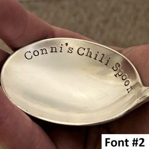May include: A silver spoon with the text "Conni's Chili Spoon" engraved on the bowl. The spoon is being held in a hand.