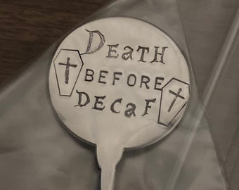 Puede incluir: Una cuchara de café plateada con una parte superior redonda, con la inscripción "Death Before Decaf" en un estilo manuscrito. El diseño incluye dos pequeños ataúdes con cruces. La cuchara está dentro de una bolsa de plástico transparente.