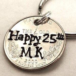 May include: A silver quarter coin keychain with the text "Happy 25th MK" stamped on it. The keychain includes a silver split ring. The coin is from the year 2000 and features the state of Maryland.