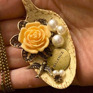 May include: A necklace with a spoon pendant featuring a yellow rose, a bird, pearls, and a charm that says "Balance".