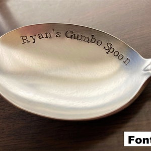 May include: A silver spoon with the text "Ryan's Gumbo Spoon" engraved on the bowl. The spoon is on a wooden surface. Font #2.