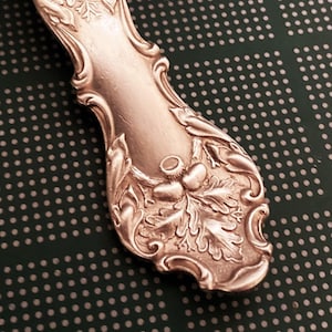May include: A silver spoon necklace with an ornate design featuring leaves and flowers. The spoon is attached to a silver chain.