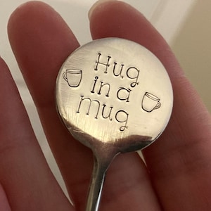 May include: A silver spoon with a round, engraved head that reads "Hug in a Mug" with a small cup graphic on either side of the text.