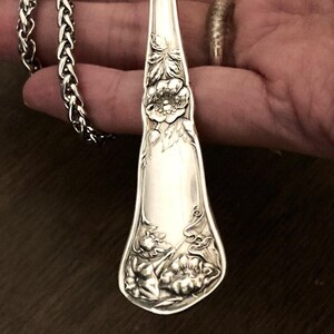 May include: A silver necklace with a long, ornate pendant made from a vintage silverware spoon. The pendant features a floral design with a large flower in the center.
