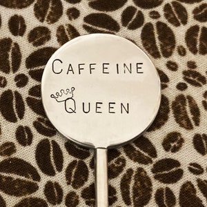May include: A silver stirring spoon with a round, flat top that says "CAFFEINE QUEEN" with a crown above the word "QUEEN". The spoon is on a brown and white fabric background with a pattern of coffee beans.