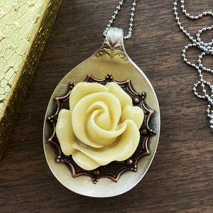 May include: A silver spoon necklace with a yellow rose charm. The rose is set in a copper-colored frame with a decorative design.